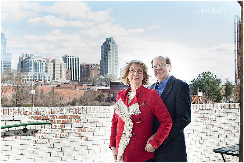 Couple standing by brick wall wearing hats and a red fringe coat with the Raleigh Sky Line