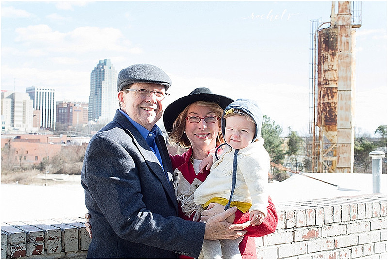 Grandparents holding granddaughter in front of a brick wall with the Raleigh Skyline behind them